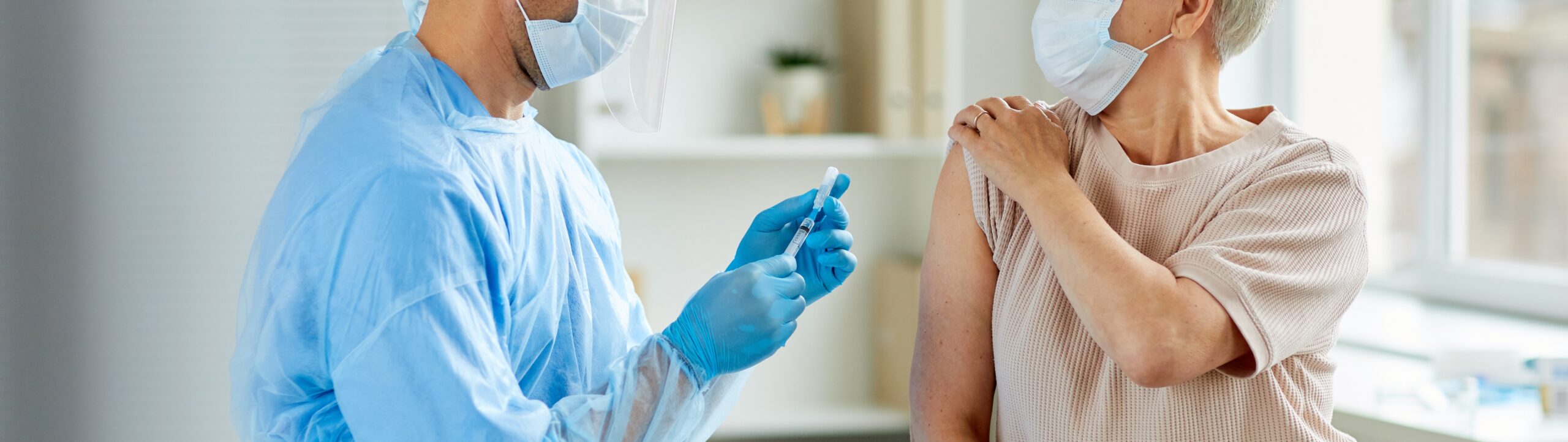 Side view shot of male nurse wearing protective mask and gloves preparing medical syringe for giving injection to senior patient
